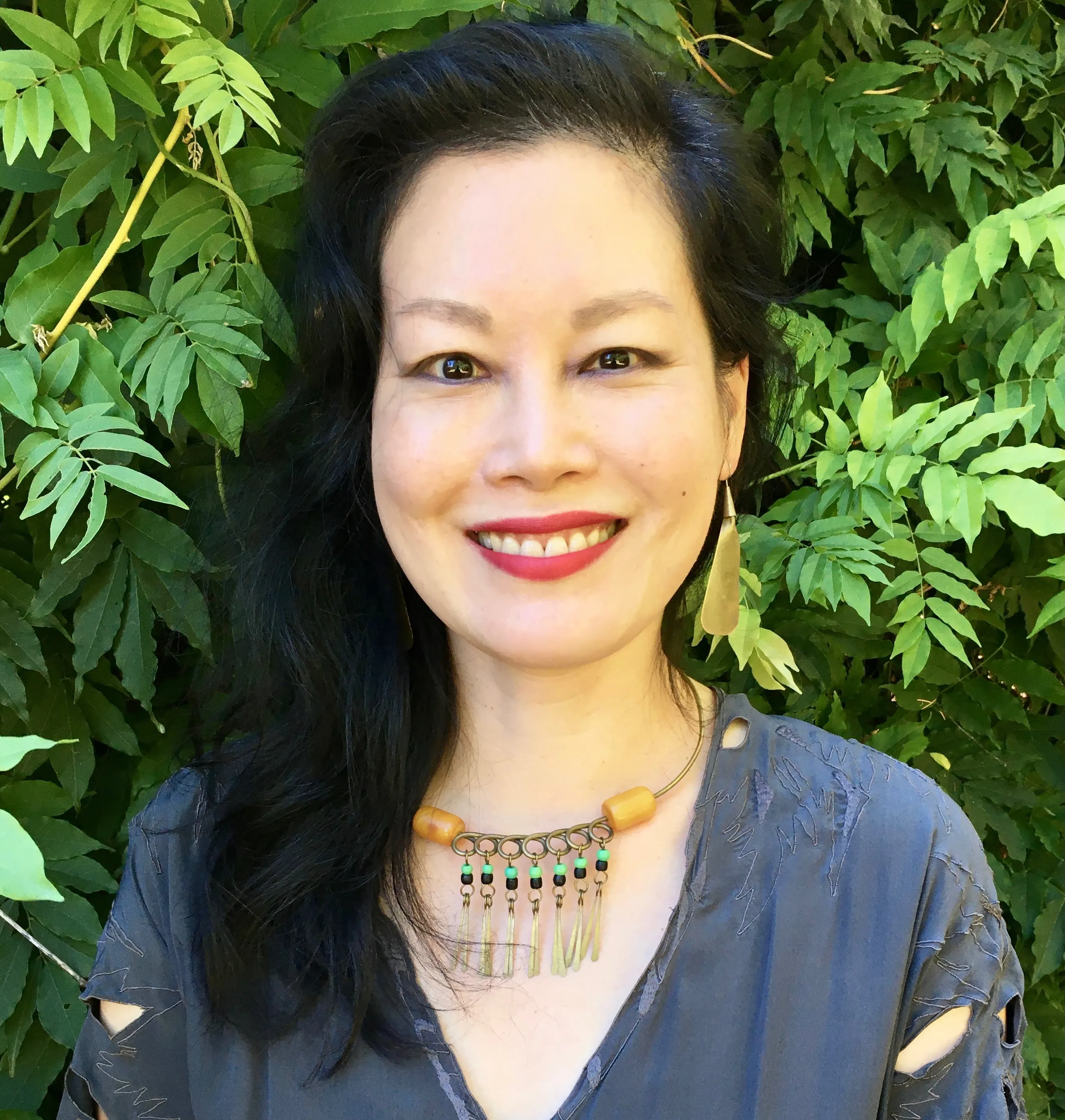 Smiling woman with long dark hair standing in front of green leafy background, wearing a blue top, a beaded necklace, and a gold earring.